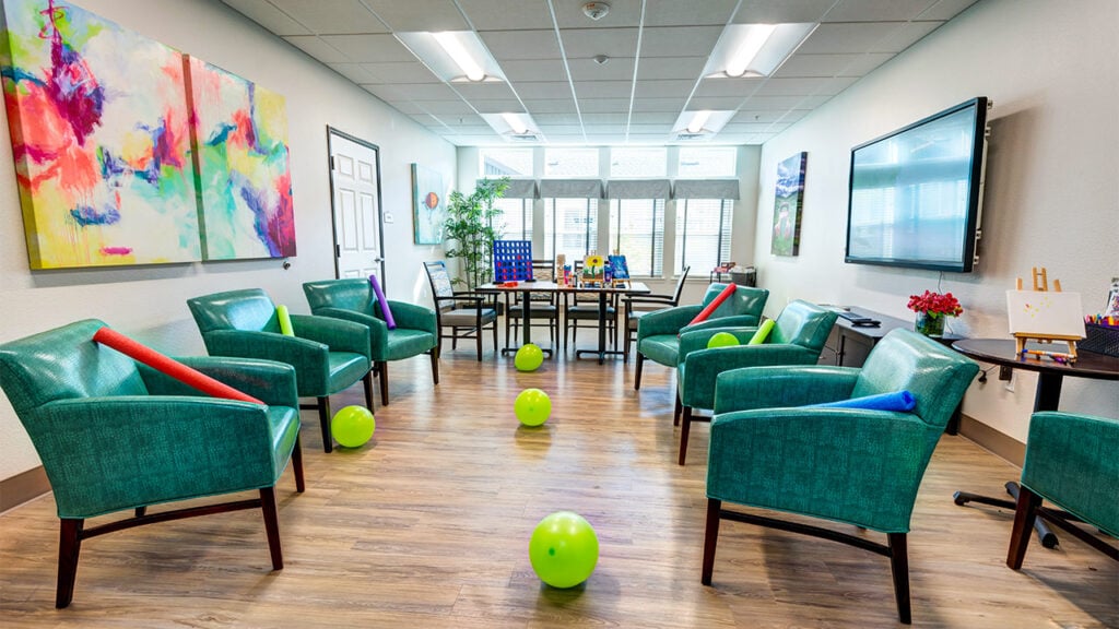 A brightly lit activity room with green chairs arranged in a circle. Colorful balloons and rolled-up blankets are on the chairs. A large TV and vibrant abstract paintings decorate the walls. Games are set up on a table near the windows.