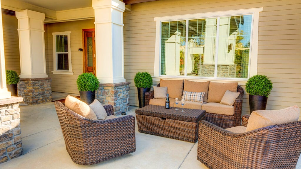 A cozy outdoor patio features a wicker furniture set with beige cushions, including two armchairs and a sofa around a rectangular table. Two glasses and a wine bottle sit on the table. Green potted plants decorate the space.