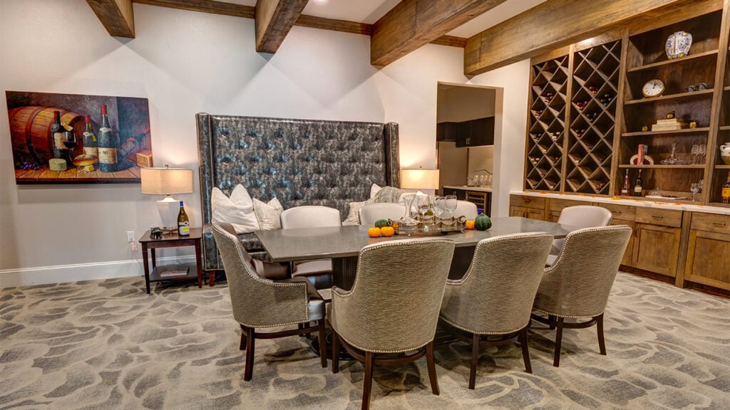 A cozy dining room with a large upholstered booth and eight cushioned chairs around a rectangular table. The room has wooden beams, a wine rack, cabinets, decorative art, and warm lighting, creating an inviting atmosphere.