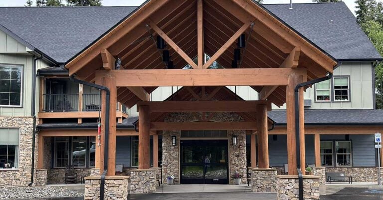 A two-story building with a wooden and stone facade. It features a peaked, timber-framed entrance with large windows and a mix of gray and beige tones. Trees and bushes are visible in the background.