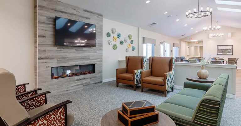 Spacious lounge with a modern, mounted TV above an electric fireplace. Two brown chairs and a green sofa face each other. Decorative wall art and round chandeliers enhance the room's style. A table with vases is set against a neutral wall.