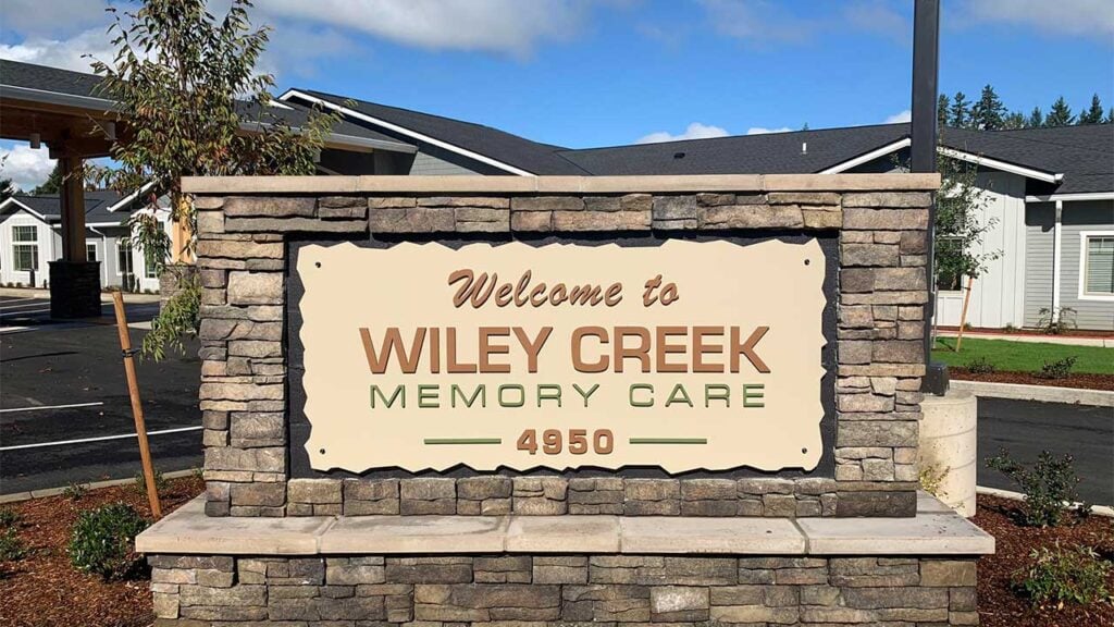 Stone sign for "Wiley Creek Memory Care" with the address 4950, set against a backdrop of residential-style buildings, trees, and a clear sky.