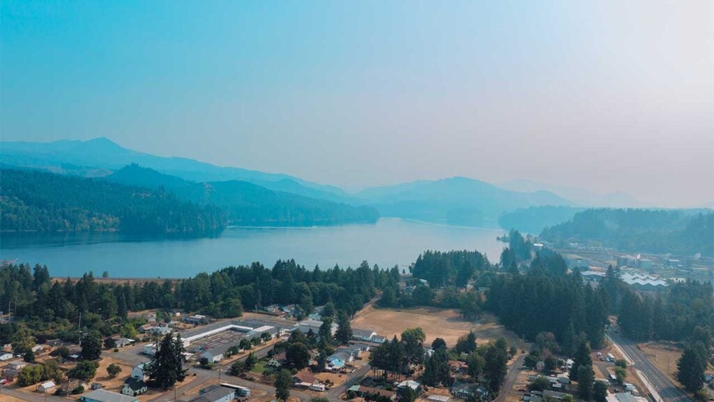 Aerial view of a serene lake surrounded by lush green forests and distant mountains under a hazy blue sky. A small town with houses and roads is visible in the foreground, blending into the natural landscape.