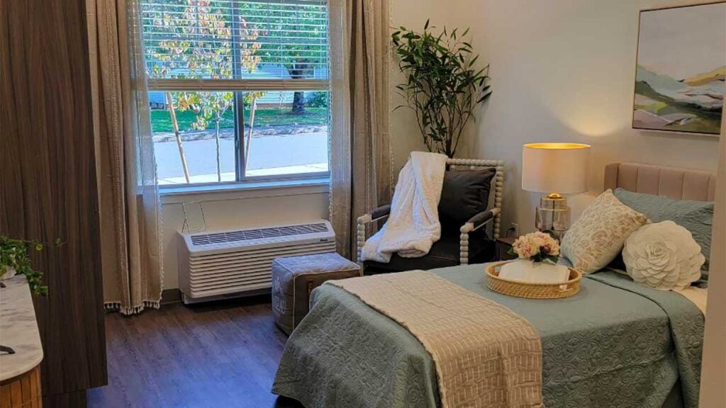 A cozy bedroom with a large window, featuring a neatly made bed with blue and beige linens, a bedside table with a lamp, and a plush chair with a white throw. A plant and an abstract painting add a touch of nature and art.