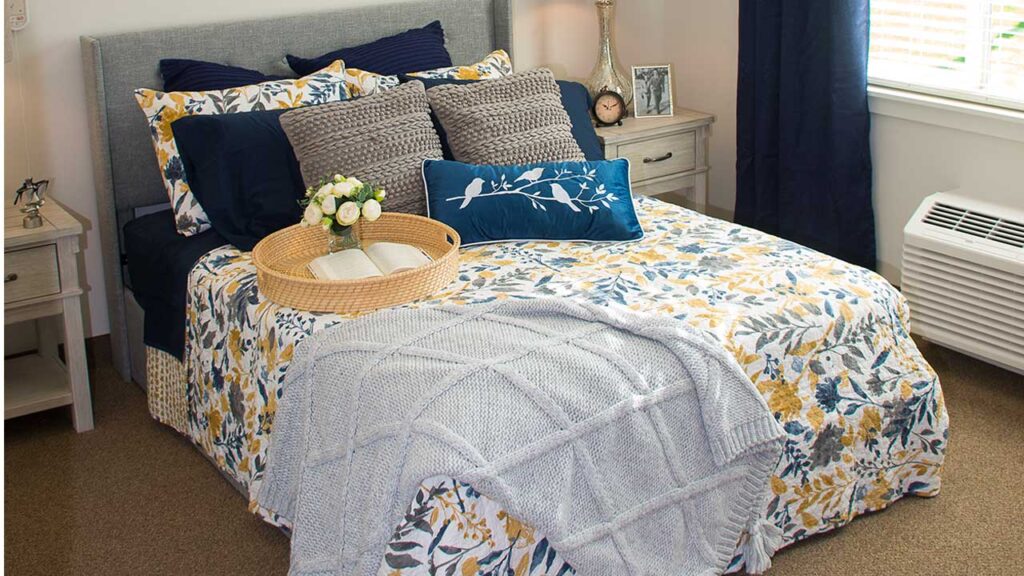 A cozy bedroom with a floral bedspread in blue, yellow, and white. The bed is adorned with pillows, a blue decorative cushion, a tray with a flower arrangement, and a folded gray blanket. Nightstands flank the bed, and a window lets in natural light.