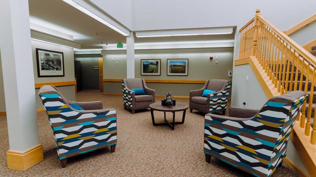 A cozy seating area in a modern hallway, featuring four patterned armchairs around a small round table. The walls are adorned with framed artwork, and a wooden staircase rises on the right. The lighting is soft and inviting.