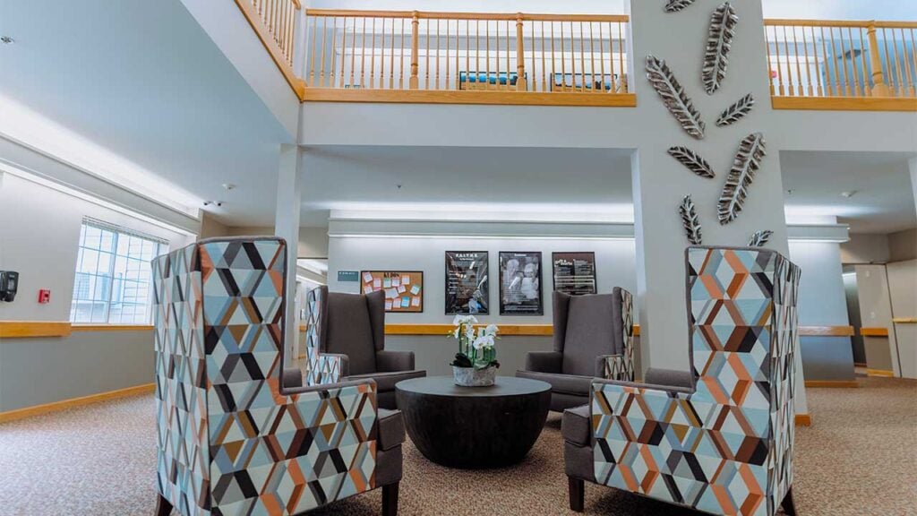 A modern lounge area with geometric-patterned armchairs surrounding a round coffee table. The room features high ceilings, large windows, and a display of decorative feathers on a central column. Two movie posters are visible on the wall in the background.