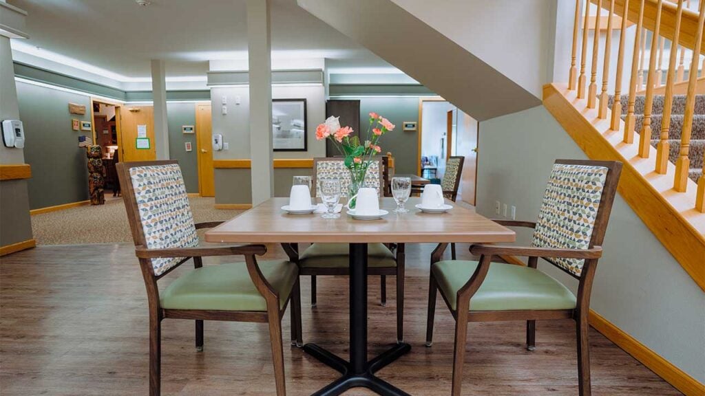 A small dining area with a wooden table set for four, featuring tea cups and a flower vase. The room has green cushioned chairs, a patterned carpet, and natural wood accents. A staircase and hallway are visible in the background.