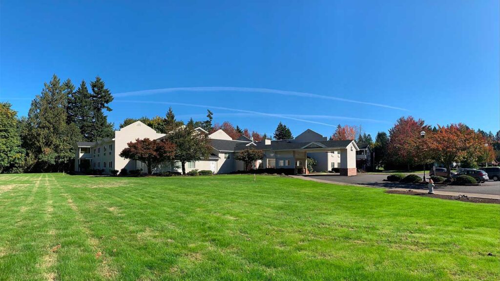 A large building with white walls and a gray roof sits at the edge of a vibrant green lawn. Trees with autumn foliage surround the building under a clear blue sky. A driveway with parked cars is visible on the right.