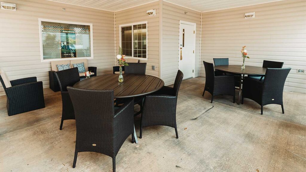 Outdoor patio with two round tables and black wicker chairs. Each table has a small vase with flowers. Beige siding and a white door are visible in the background.