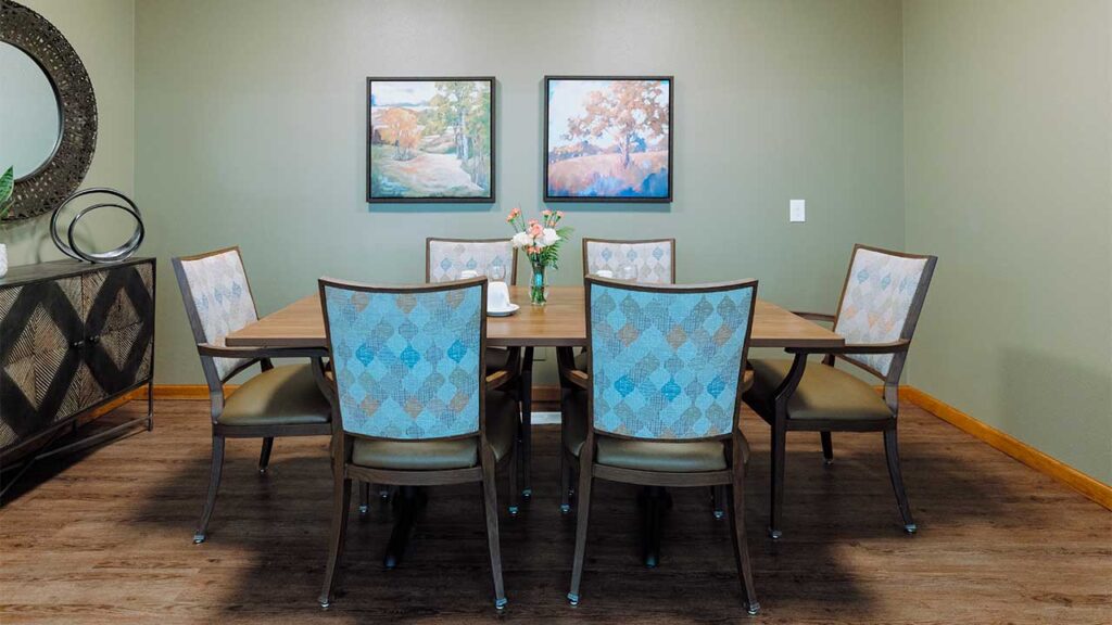 A dining room features a wooden table with six upholstered chairs. Two landscape paintings hang on the green wall. A sideboard with a round mirror is on the left, and a vase with flowers sits on the table. The floor is wooden.