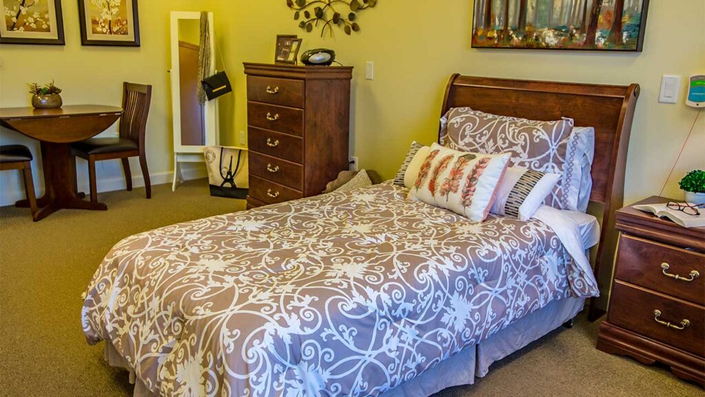 A cozy bedroom with a wooden bed featuring patterned bedding and decorative pillows. A nightstand with a clock and picture frames is beside the bed. A small dining table with two chairs and wall art complete the warm decor.