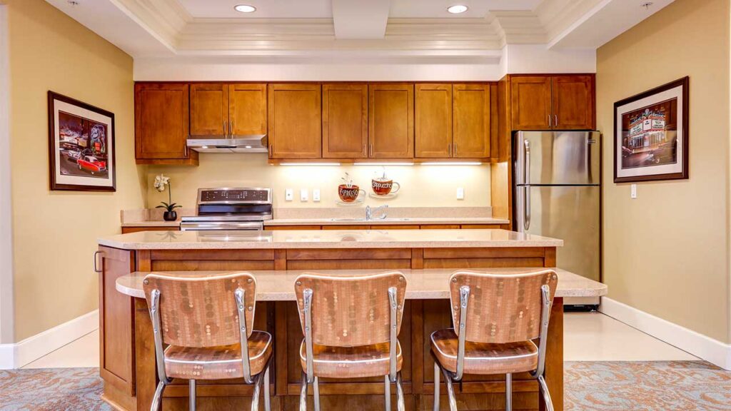 A kitchen with wooden cabinets and a matching island. Three brown stools are placed at the island. The space includes a stainless steel refrigerator, an oven with a hood, and wall art on either side. The room is lit with ceiling lights.