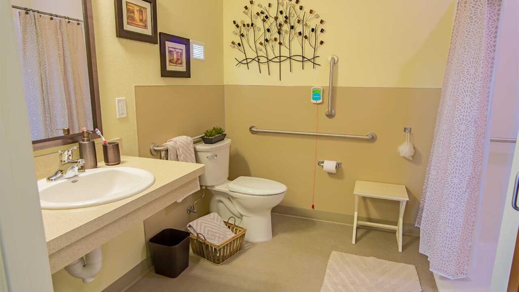 Accessible bathroom with a wall-mounted sink, grab bars near the toilet, a decorative wall piece, and a shower with a curtain. The room has neutral-colored walls and flooring, a small stool, and a woven basket with towels.