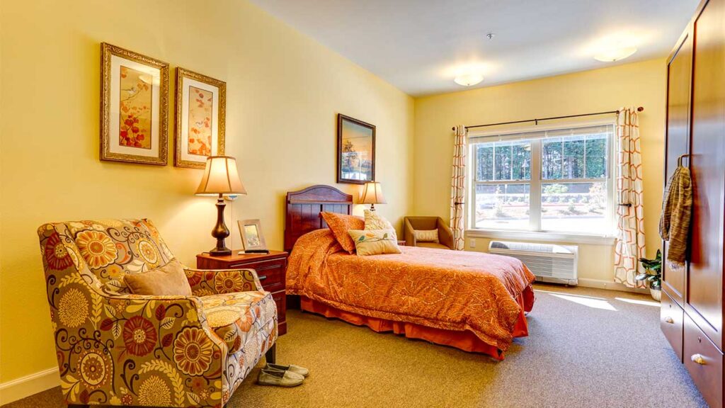 Cozy bedroom with a floral armchair, an orange-patterned bedspread, wooden nightstand with lamp, and framed art on pale yellow walls. Sunlight streams in through a large window with checkered curtains, illuminating the warm, inviting space.