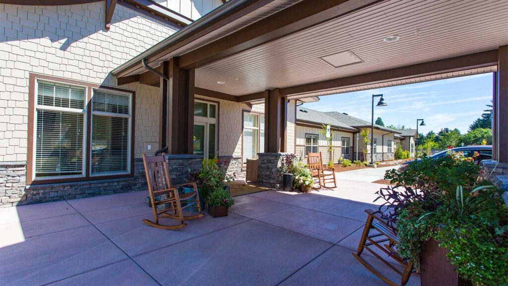 Covered patio with rocking chairs and potted plants outside a building. Exterior features stone accents and large windows. The patio is adjacent to a parking area with lampposts and greenery in the background.