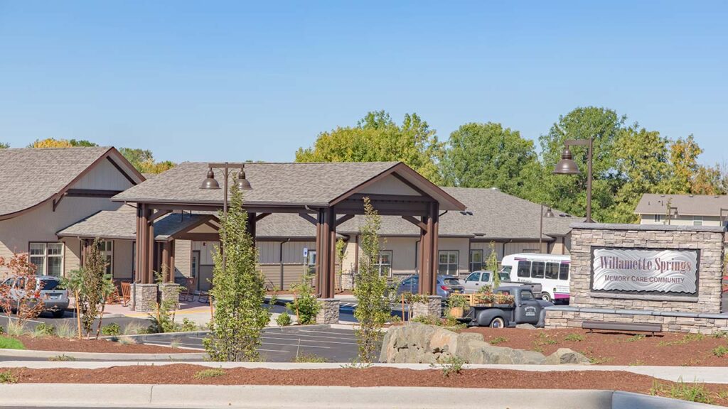 A large wooden building with a covered entrance and a manicured garden. A sign reads "Willamette Springs Memory Care Community" next to the driveway. Trees in the background under a clear blue sky.