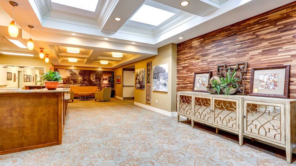 A warmly lit, spacious lobby area featuring a wooden reception desk, contemporary art, and a decorative wood-accented wall. Comfortable seating with orange chairs is visible in the background. Skylights and pendant lights enhance the bright, welcoming atmosphere.