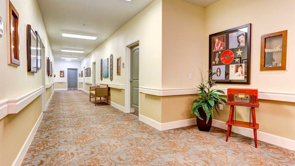 A well-lit hallway with framed photos and memorabilia on the walls, a plant in the corner, and a vintage wooden radio on a stand. The carpet has a subtle floral pattern, and doors line one side of the corridor.