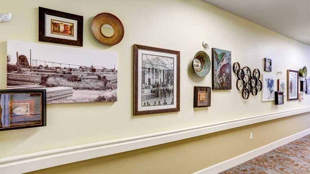 Hallway with a gallery wall displaying an eclectic mix of framed photos, art, and decorative objects, including circular metal sculptures and vintage photographs. The arrangement is on a light-colored wall with a patterned carpet below.