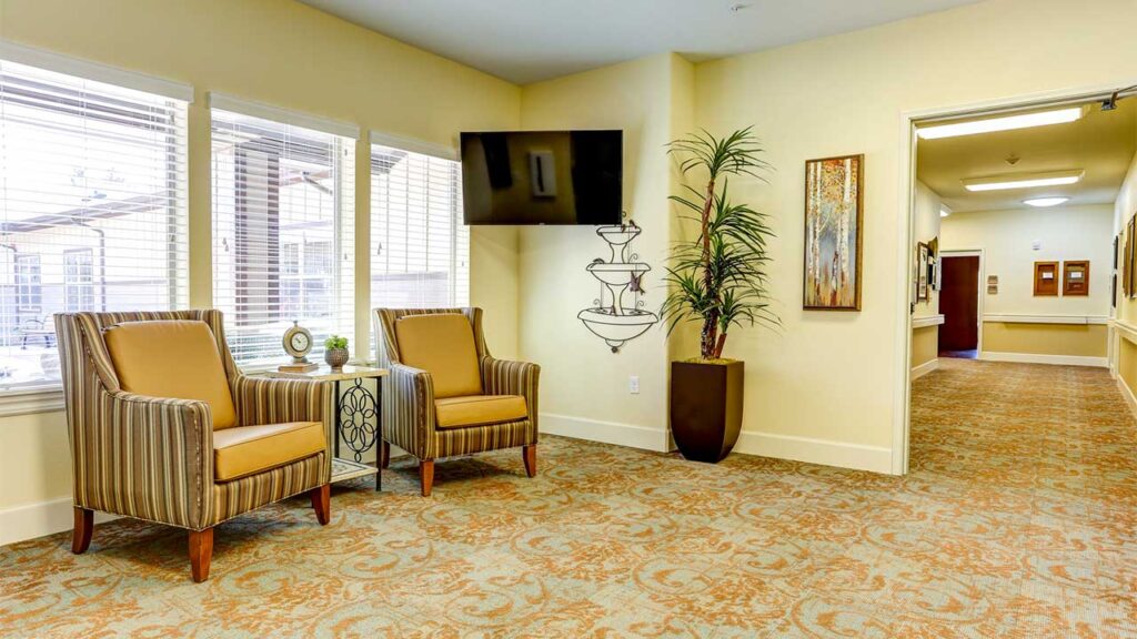 A cozy waiting area with two upholstered chairs and a small table by large windows, featuring a flat-screen TV and a decorative potted plant. A hallway extends to the right, adorned with framed art on the walls and patterned carpeting.