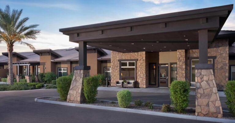 A modern, single-story building with stone facade and dark wooden accents. The entrance is sheltered by a large awning supported by stone pillars. The surrounding landscaping features small bushes and a palm tree under a clear blue sky.