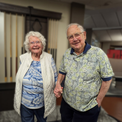 An elderly couple stands indoors, both smiling. The woman is wearing a white quilted vest over a blue patterned shirt, and the man is in a floral shirt. They are holding hands, with a blurred background of a room.