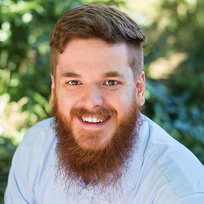 A bearded man with brown hair is smiling and wearing a light blue shirt. He is outdoors with greenery in the blurred background.