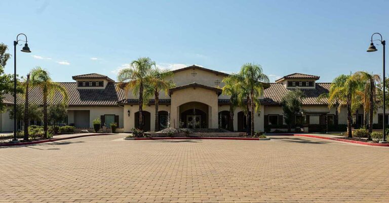 A large Spanish-style building with a beige stucco exterior and a terracotta tile roof. The front entrance is flanked by several tall palm trees, and there are lamp posts lining the driveway. The sky is clear and blue.