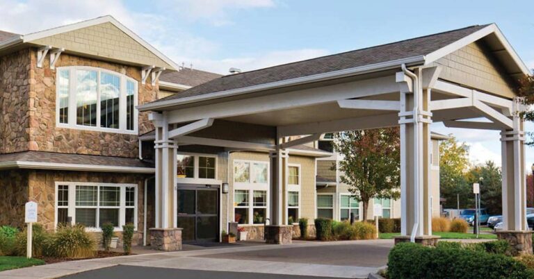 A modern building with stone and siding exterior features a covered entrance supported by white columns. Large windows and neatly landscaped grounds with trees and shrubs surround the structure.