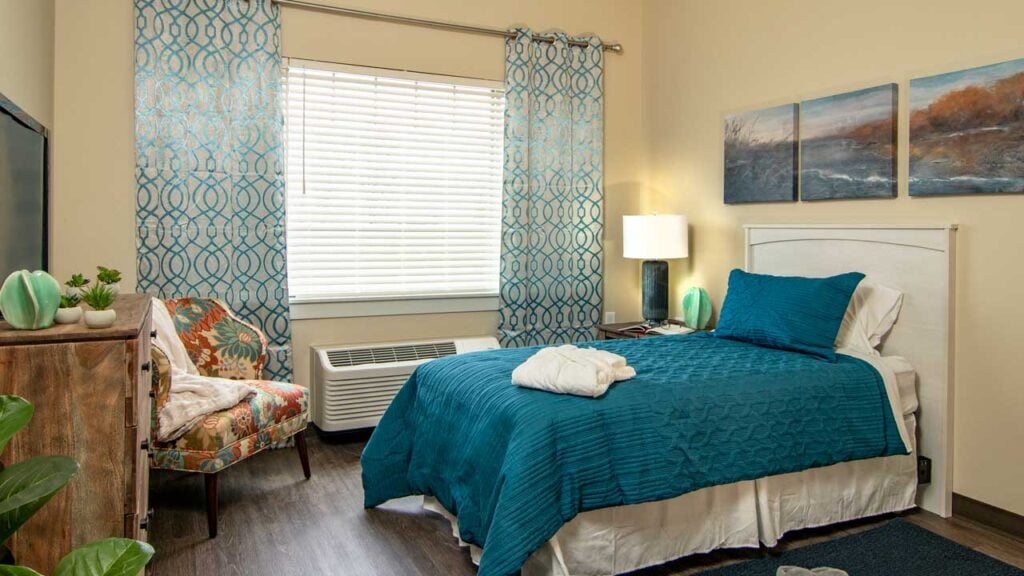 A cozy bedroom with a neatly made bed featuring teal bedding. The room has patterned curtains, a trio of landscape paintings above the bed, a floral chair, and a wooden dresser. A window with blinds lets in natural light.