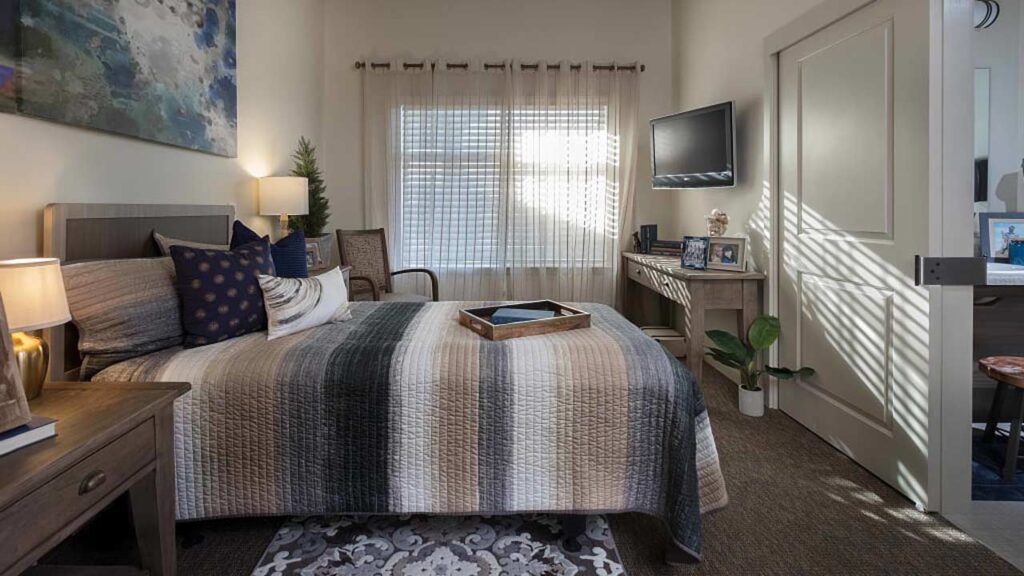 A cozy bedroom with a striped bedspread, multiple pillows, and a wooden headboard. A small lamp on the nightstand illuminates the room. A television is mounted on the wall, and a tray rests on the bed. Sheer curtains cover the window.