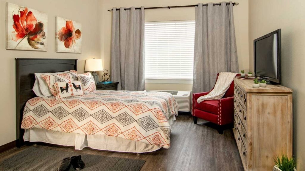 Cozy bedroom with a queen-sized bed featuring patterned bedding. Red flowers adorn the wall above. A red armchair sits near a wooden dresser with a TV. Grey curtains cover the window, and a lamp softly lights the room.