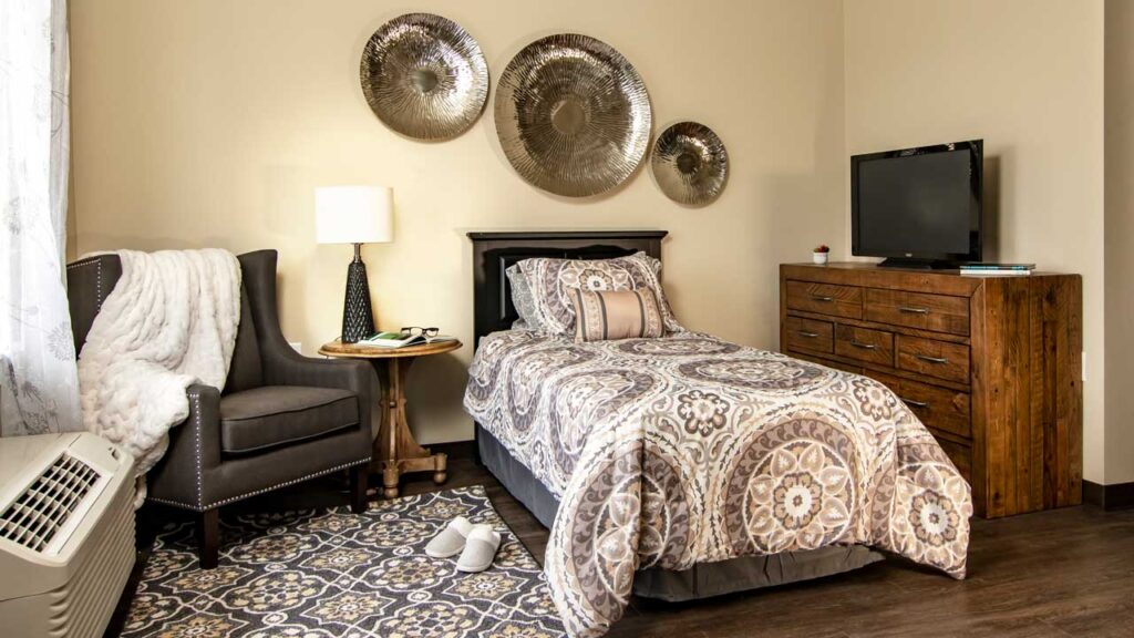 A cozy bedroom with a twin bed featuring a patterned comforter, a wooden dresser with a TV, and a gray armchair beside a small round table. Decorative metal wall art is displayed above the bed, and a soft rug covers the floor.