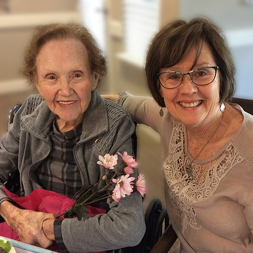 Two smiling women sitting together indoors. The older woman on the left is holding pink flowers, while the other woman rests her arm around her. Both are wearing glasses and casual clothing. The background is softly blurred.