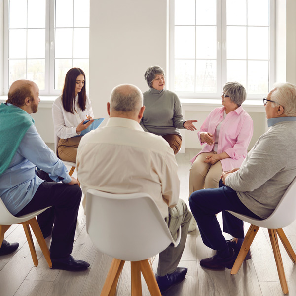 A group of older adults sits in a circle on chairs, engaged in a discussion. A younger woman with a clipboard listens and takes notes. Large windows provide natural light in the bright room.