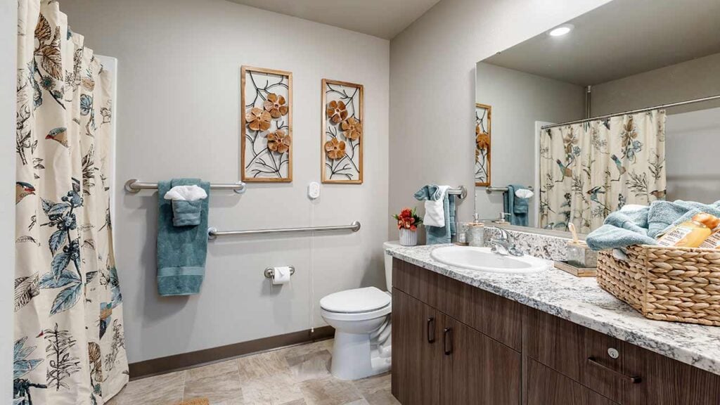 A modern bathroom features a shower with nature-themed curtains, a toilet with grab bars, a large mirror above a wooden vanity, and a basket of towels on the counter. Wall art depicting flowers adds a decorative touch.