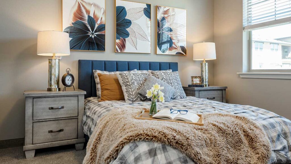 Cozy bedroom with a neatly made bed featuring patterned pillows, a plush blanket, and a tray with glasses and a book. Nightstands with lamps flank the bed. Three floral artworks hang above, and sunlight streams through a window.