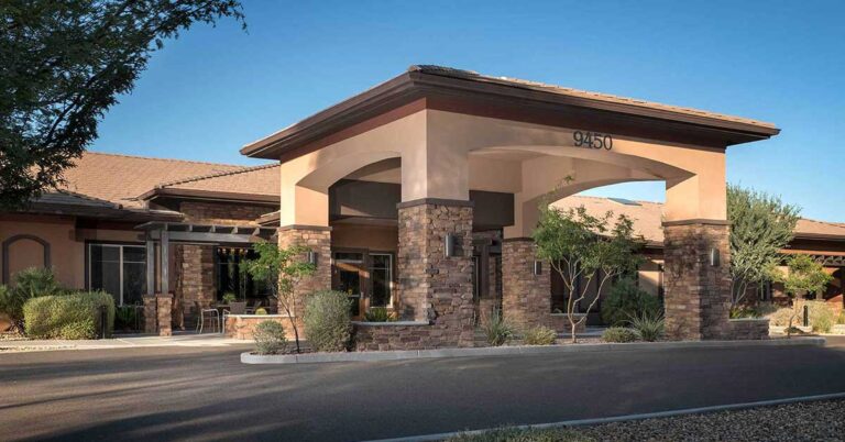 A modern, single-story building with stone and stucco exterior, featuring a covered entrance. Surrounded by landscaping with small trees and shrubs, the structure is set against a clear blue sky. The number 9450 is displayed above the entrance.