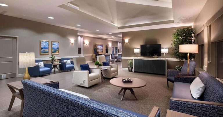A modern, spacious living room with comfortable seating, including sofas and armchairs. The room has warm lighting, a flat-screen TV, decorative plants, and artwork on the walls, creating an inviting and cozy atmosphere.