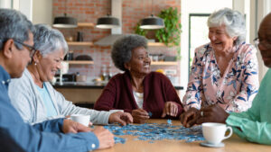 Five older adults sit around a table, smiling and laughing while working on a jigsaw puzzle together in a cozy, modern kitchen—a heartwarming glimpse into daily life that answers the question, “What is assisted living?” Two coffee cups rest on the table, emphasizing the warmth and comfort these supportive communities provide.