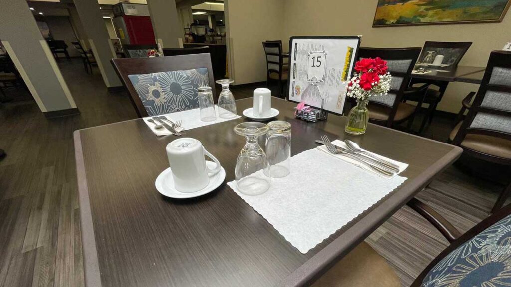 A restaurant table set for two with white placemats, glasses, cutlery, and white napkins. A vase with red and white flowers and a table number card are placed near the center. Wooden chairs and other tables are visible in the background.