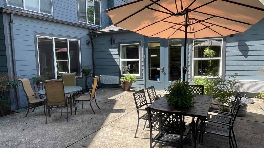 A sunny outdoor patio with a round table and chairs on the left, and a square table with chairs on the right under a large umbrella. The area is surrounded by blue-gray walls with windows, and there are potted plants around.