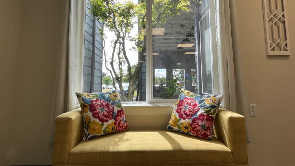 A cozy room with a light yellow couch featuring two floral-patterned pillows. Large window behind the couch provides natural light, revealing a view of trees and a building exterior. Walls are light-colored, creating a serene atmosphere.