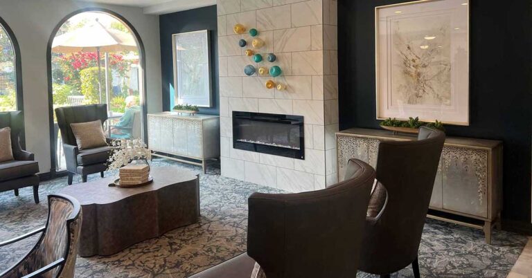 A modern living room with a central fireplace surrounded by marble. Two dark armchairs face a coffee table. Large artwork hangs on the walls, and arched windows offer a view of a garden outside. Decor includes a wall-mounted glass sculpture.