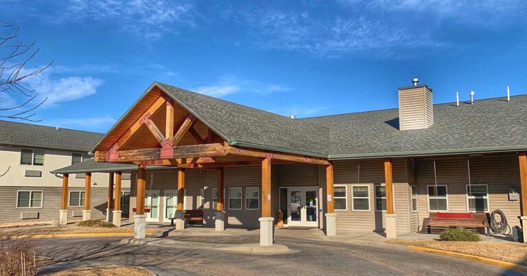 A single-story building with a wooden entrance canopy and pitched roof on a sunny day. The facade is gray with large windows, and there are shrubs and a paved walkway in front. A red bench is visible to the right of the entrance.