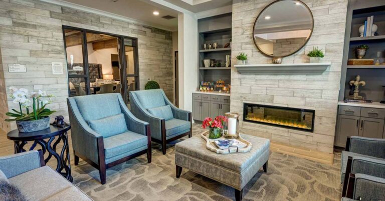 A cozy living room with light gray walls and a large fireplace. Two blue armchairs and a gray ottoman are centered on a patterned rug. Shelves flank the fireplace, and a round mirror hangs above. A glass door leads to another room.