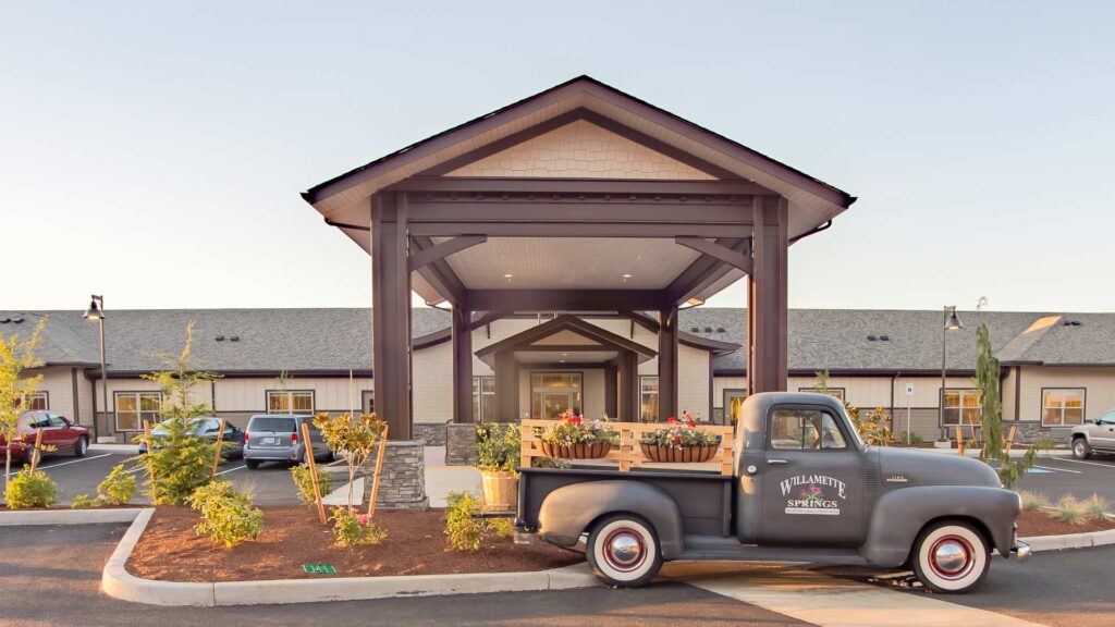 Front view of a building with a peaked roof and an entrance walkway. An old gray truck with "Willamette Springs" on the door is parked in front. The surrounding area features landscaped gardens and parked cars.