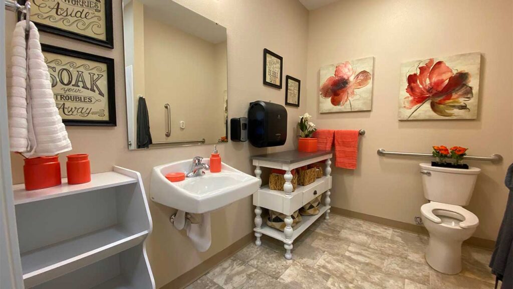 A bathroom with a white sink and toilet, decorated with red and orange accents. Floral artworks adorn the walls, and there are towels and a small shelf with baskets. The space includes a large mirror and a wall-mounted hand towel rack.