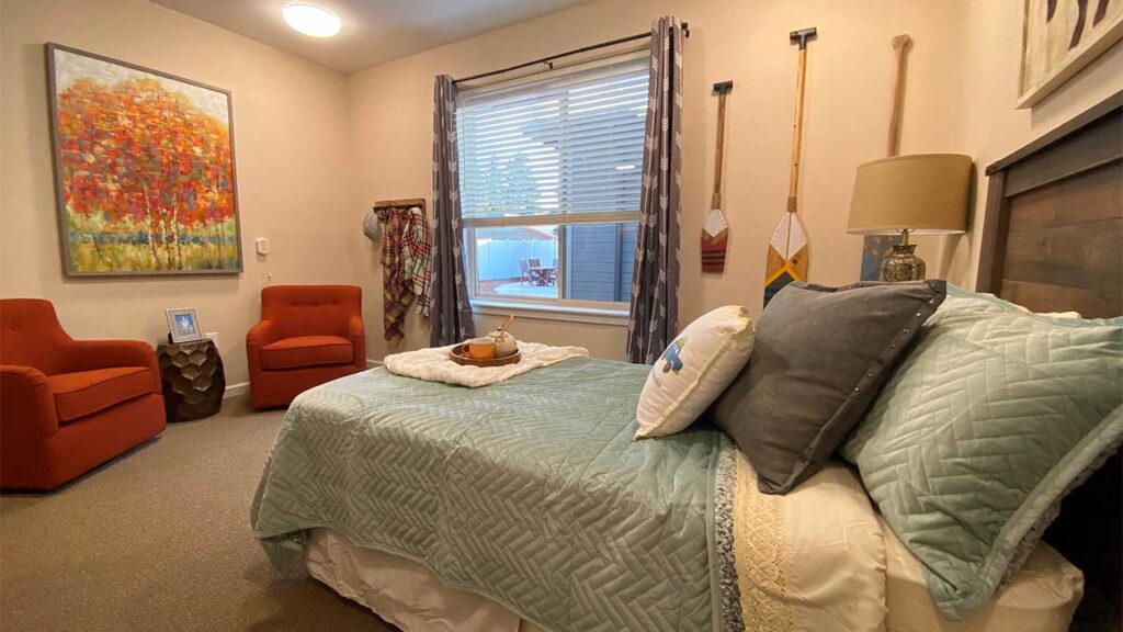 A cozy bedroom with a neatly made bed covered in teal and white linens. Two orange chairs sit near a colorful painting of trees. A window with blinds lets in natural light, and decorative oars hang on the wall. A tray with items rests on the bed.